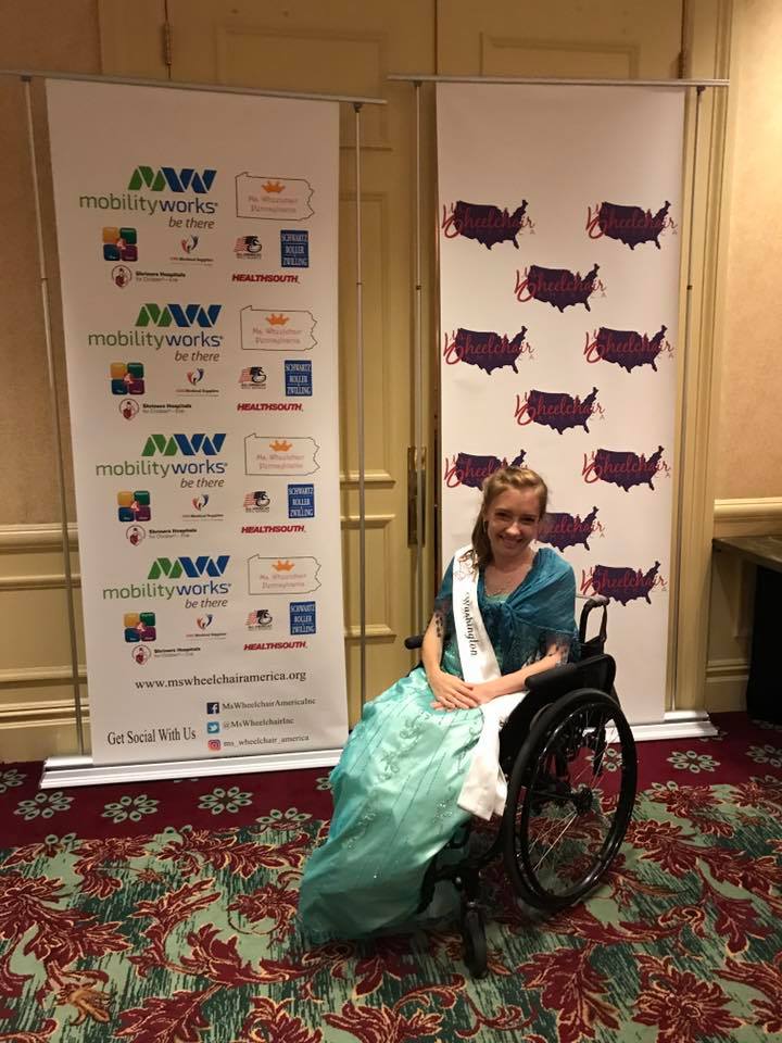 Kyann is sitting in her chair with her hands folded on her lap. She is sitting in front of the Ms. Wheelchair America sign.