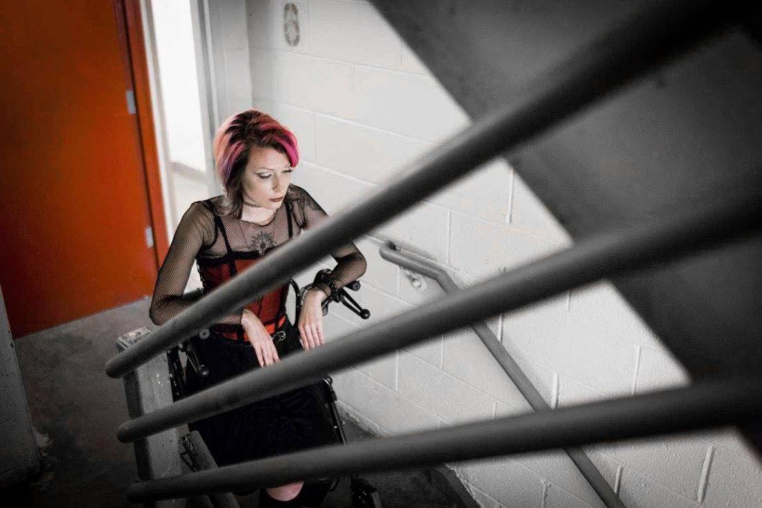 Anomie sits in her powerchair at the top of a set of stairs in a building stairwell, looking down at the steps she cannot use with her chair.